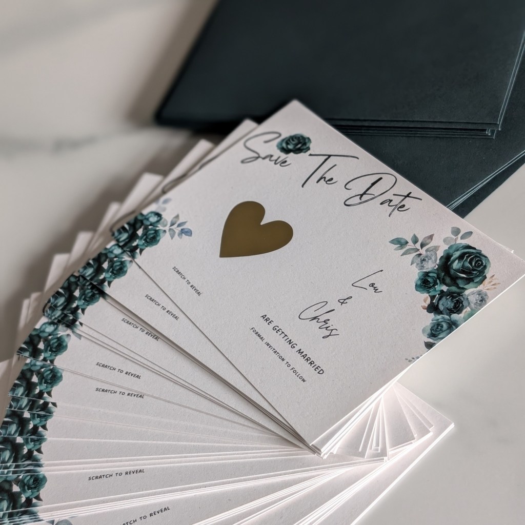 A stack of 'Save The Date' wedding cards featuring floral designs and a gold heart scratchcard sticker, with matching envelopes in the background.